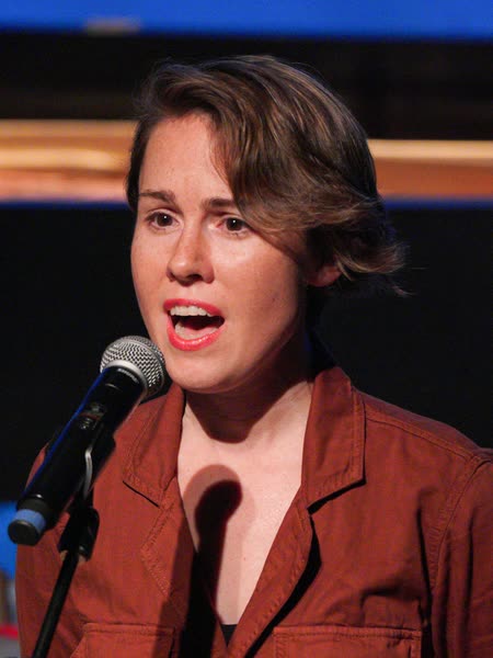 Color photograph of Caroline Shaw singing into a microphone on stage, wearing a rust-colored shirt