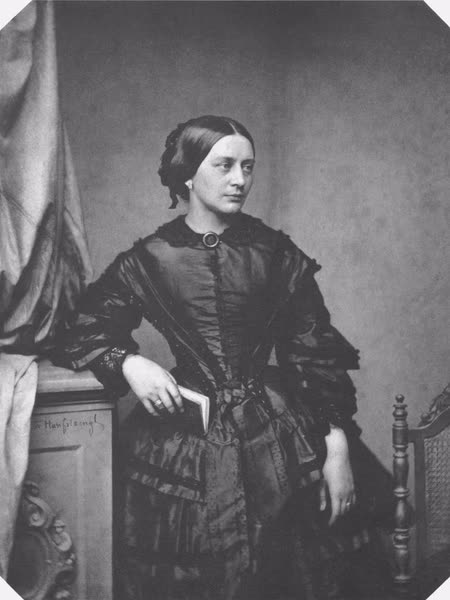 Photograph of Clara Schumann standing in a dark silk dress, her hair parted in the middle, resting one hand on a table