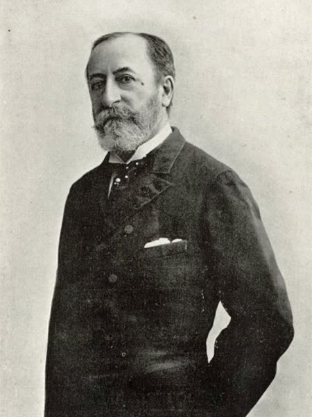 Photograph of Camille Saint-Saëns standing in a dark frock coat, with a short beard, one hand in his pocket